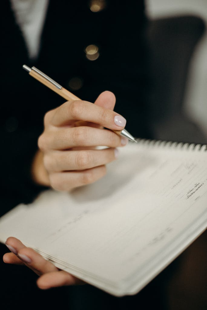 A close-up of a hand holding a pen and writing in a spiral notebook.